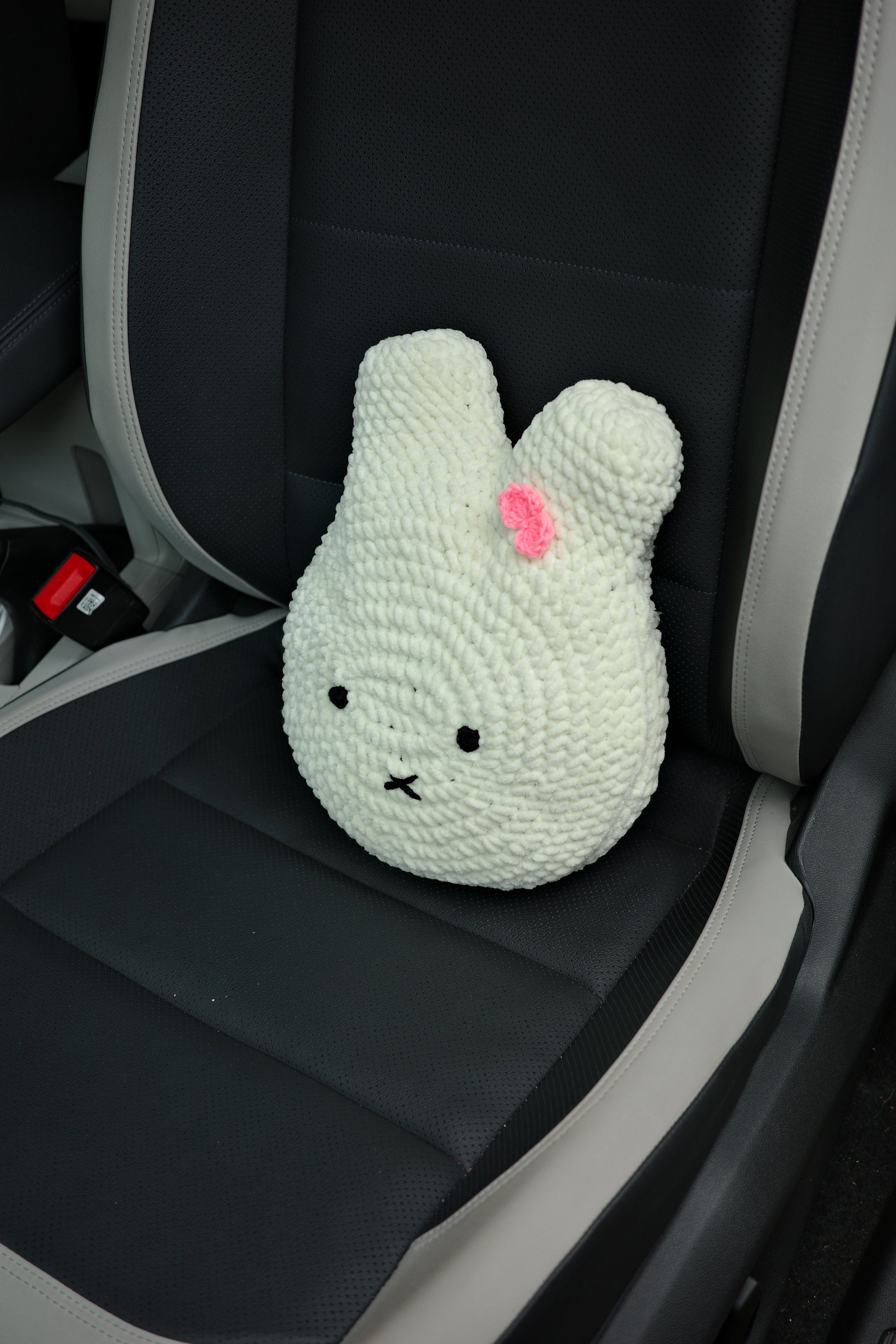 White knitted miffy cushion toy with black eyes and a pink heart on a car seat.