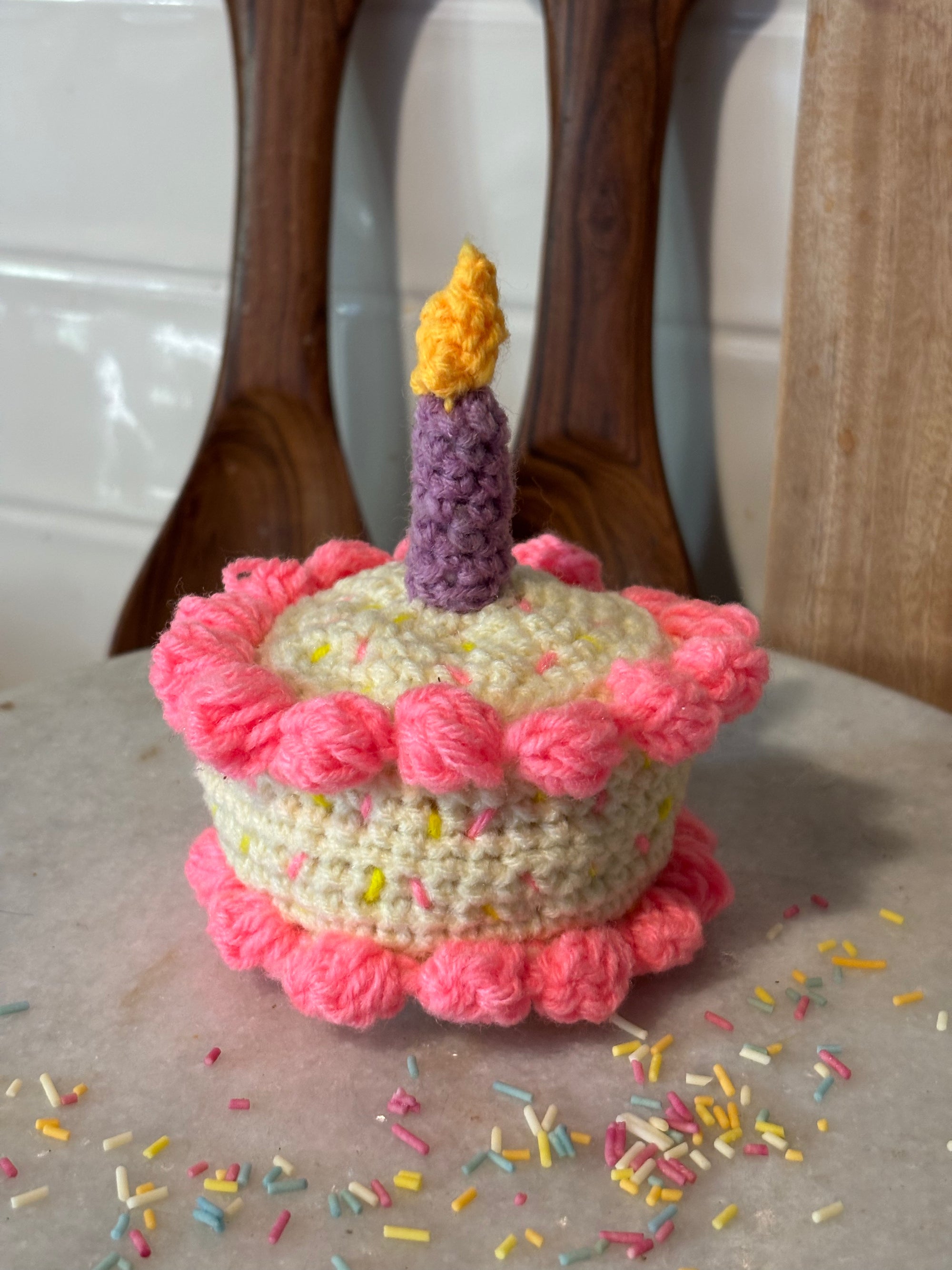 Crocheted birthday cake with pink frosting and a single candle on a surface with scattered sprinkles.