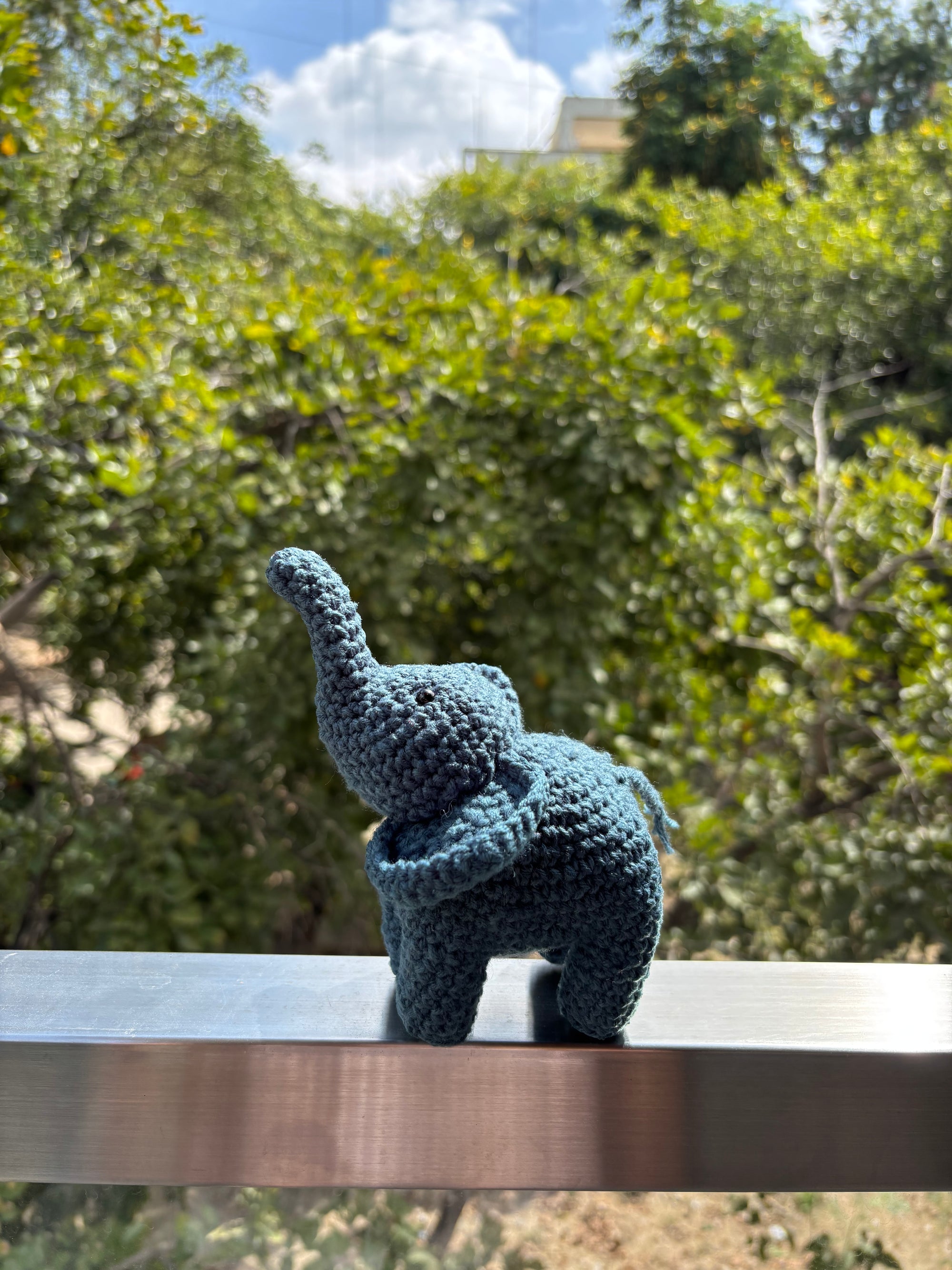 Blue grey knitted elephant toy on a metal surface with greenery in the background