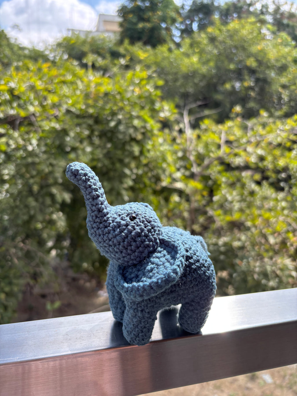 Blue grey crochet elephant toy on a wooden surface with greenery in the background