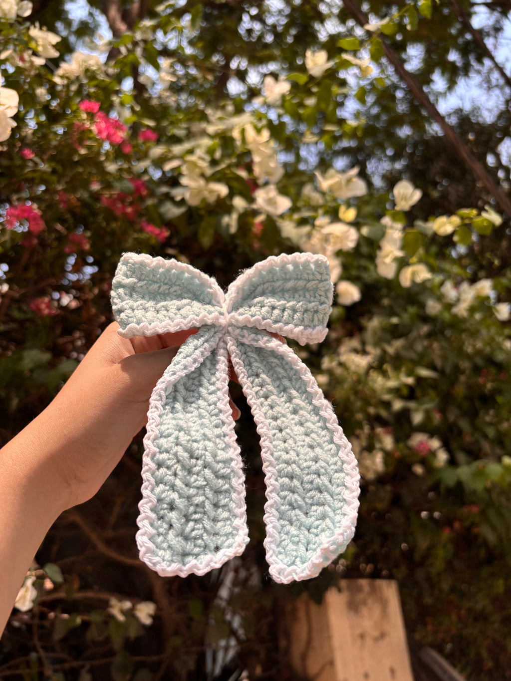 Hand holding a light blue crocheted bow with a blurred floral background