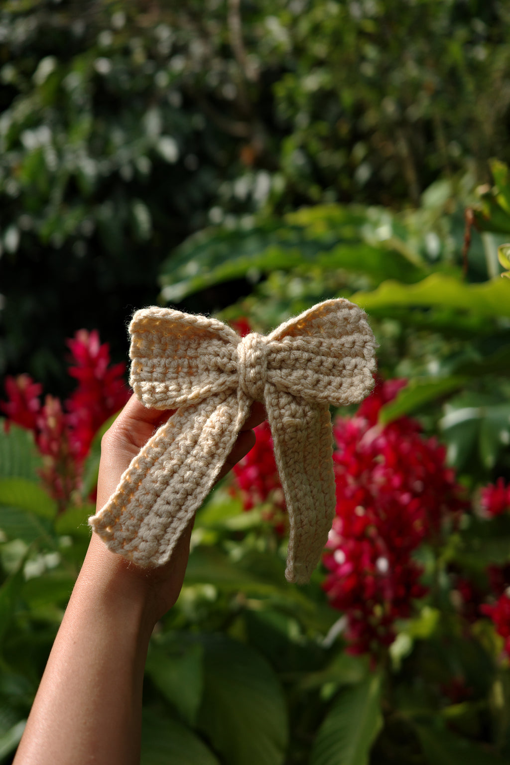 Hand holding a beige crochet butterfly against a blurred natural background with red flowers.