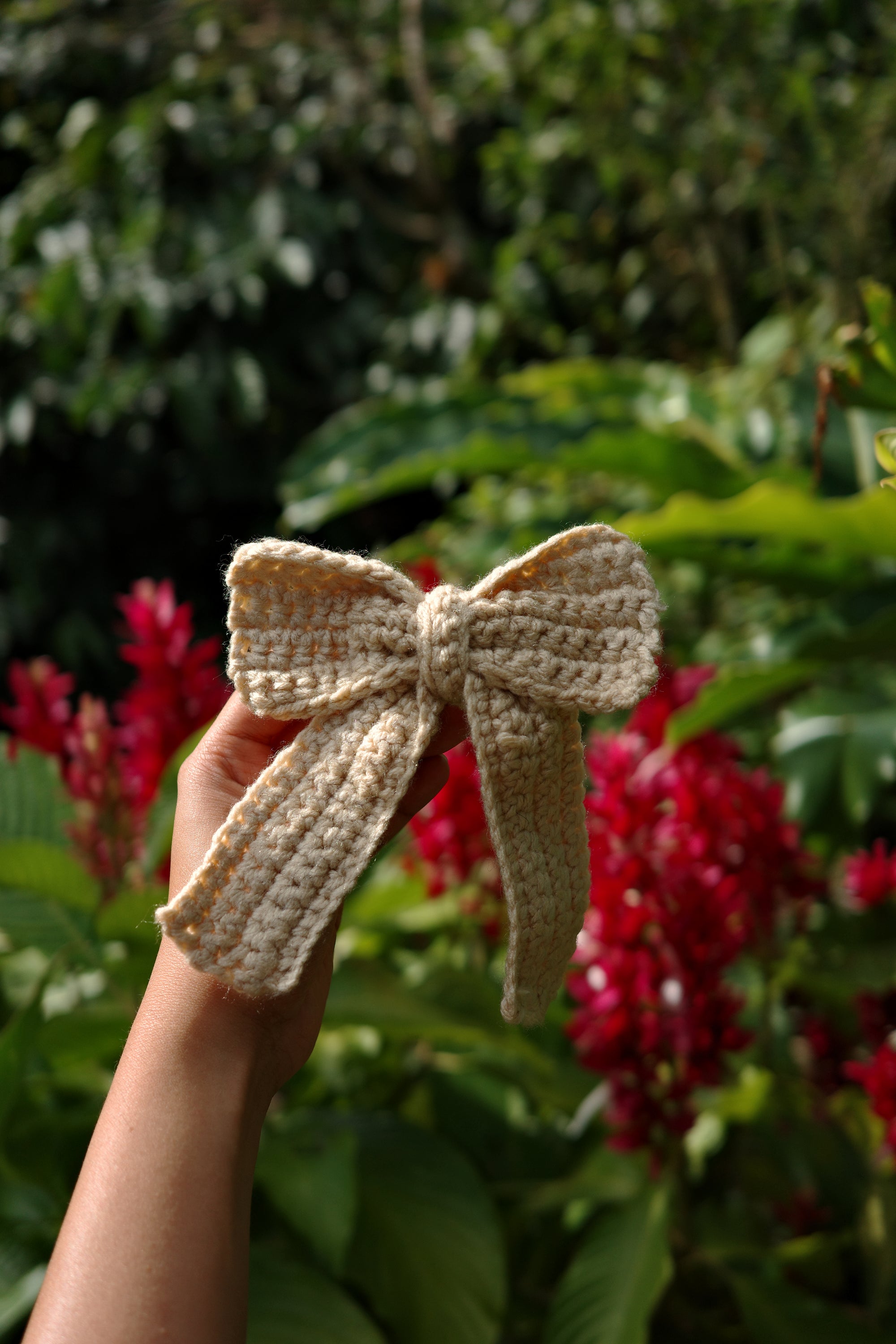 Hand holding a beige crochet butterfly against a blurred natural background with red flowers.