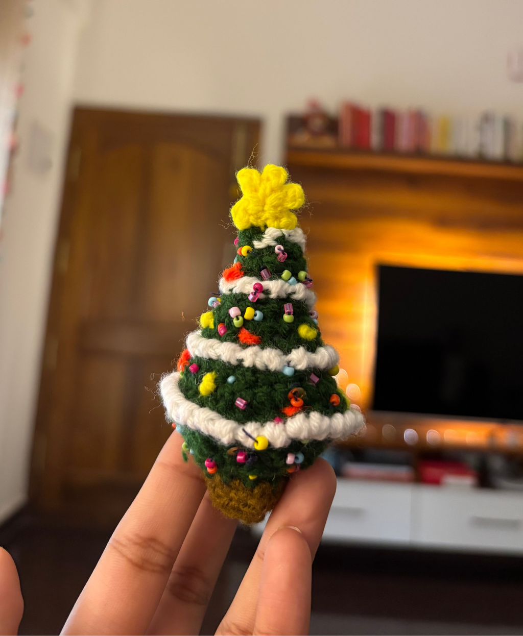 Hand holding a small crocheted Christmas tree with colorful decorations in a room.