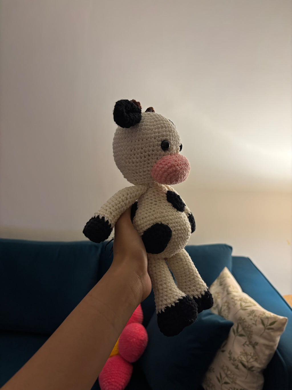 Hand holding a crochet amigurami cow toy against a blue couch and light-colored wall.