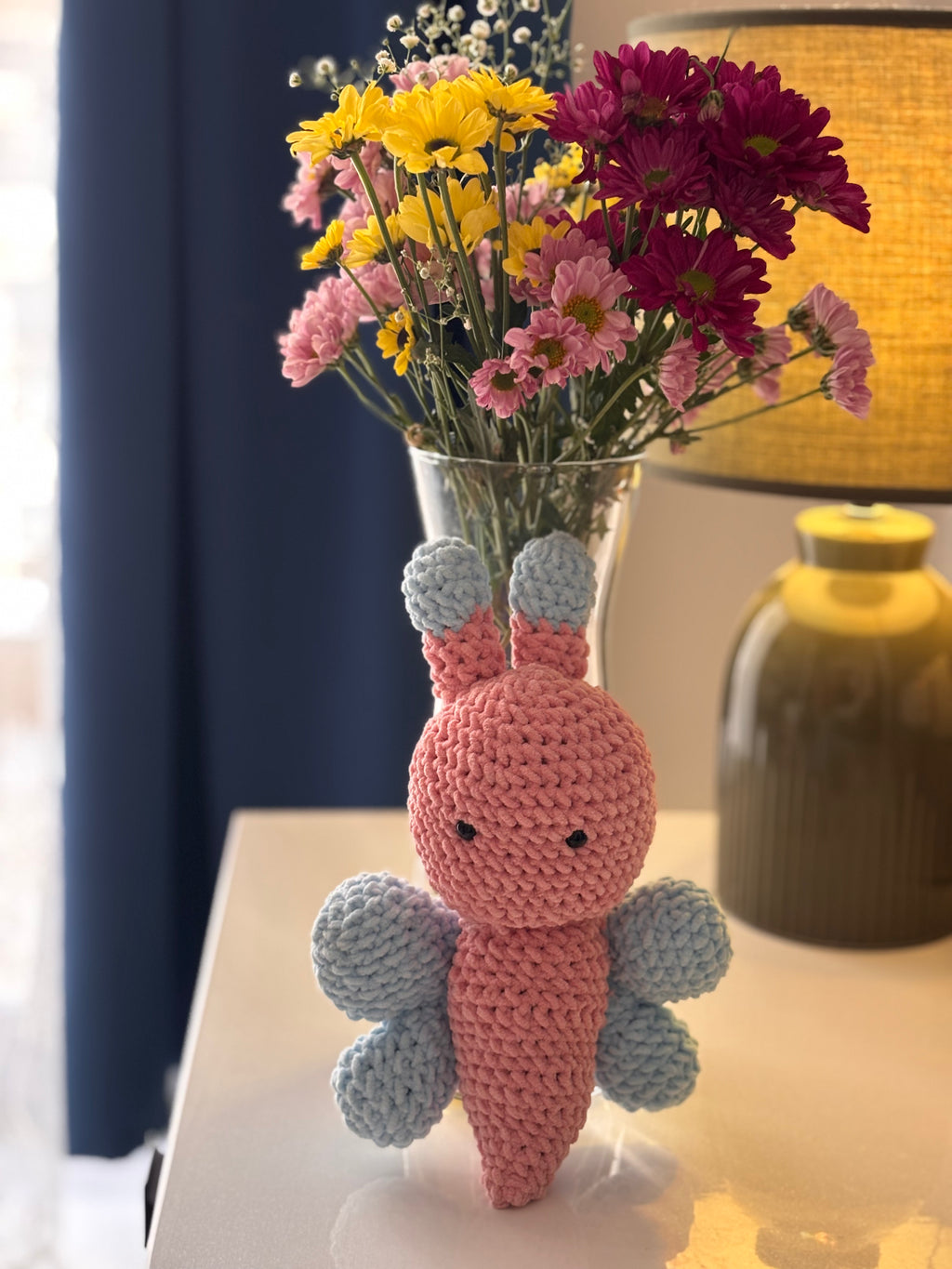 Crocheted butterfly figure with a glass vase of flowers on a table.