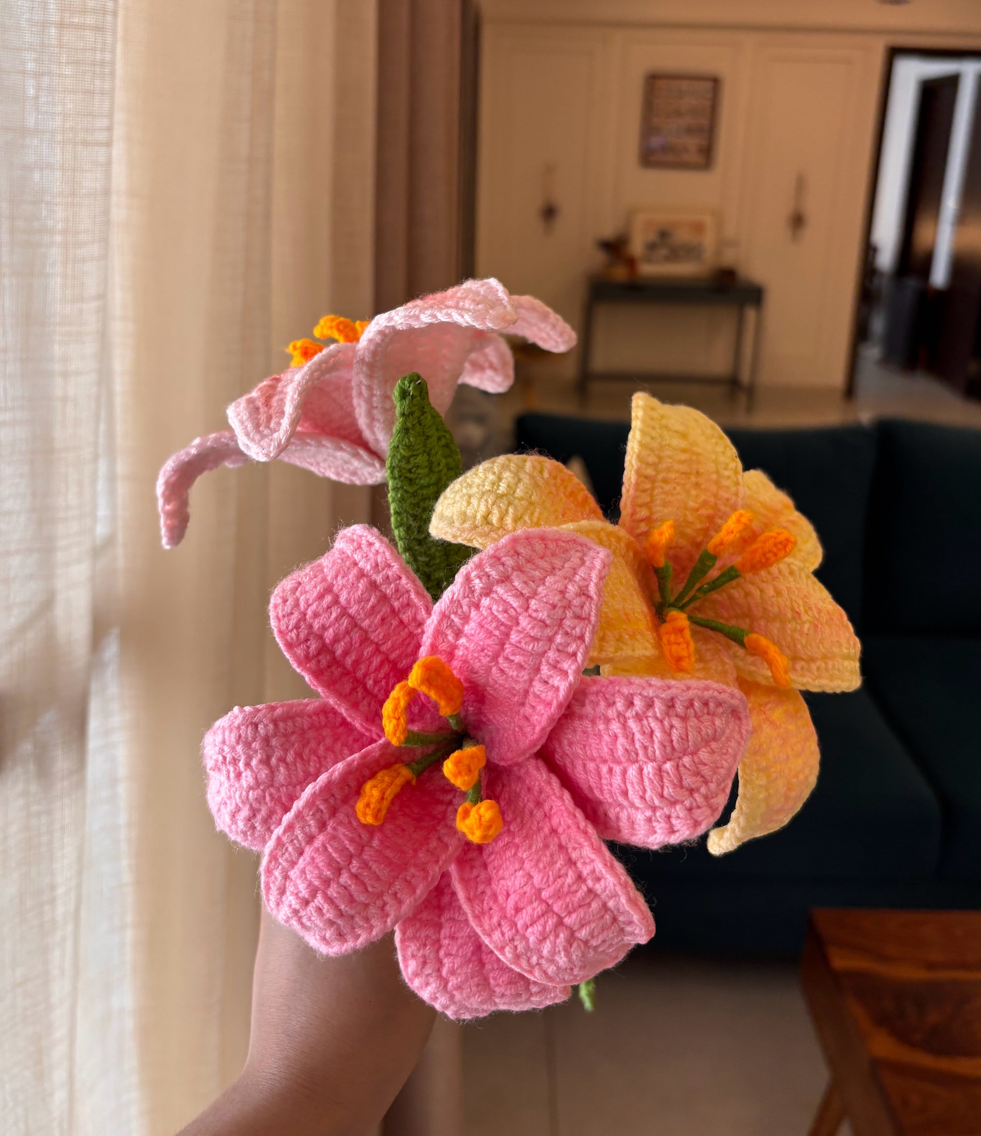 A hand holding a handmade crochet bouquet of three lily-like flowers in pink, yellow, and peach with orange centers and green stems, set against a blurred living room background.