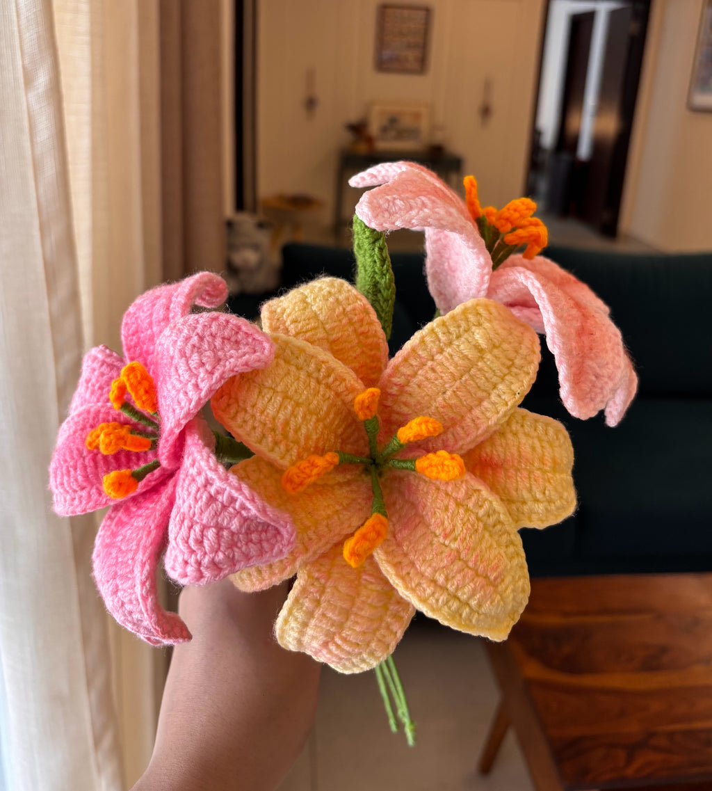 A hand holding three crocheted lily flowers in pink and pale yellow with orange centers, photographed indoors with a blurred living room background.