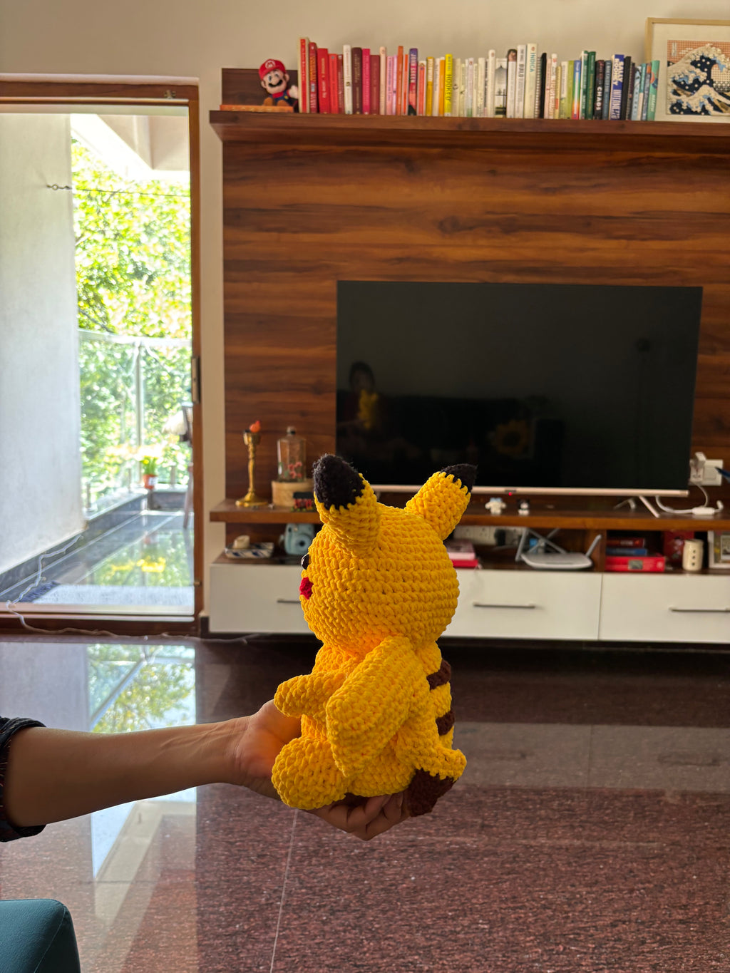 Hand holding a yellow knitted Pikachu crochet toy in a living room with a fireplace and books.