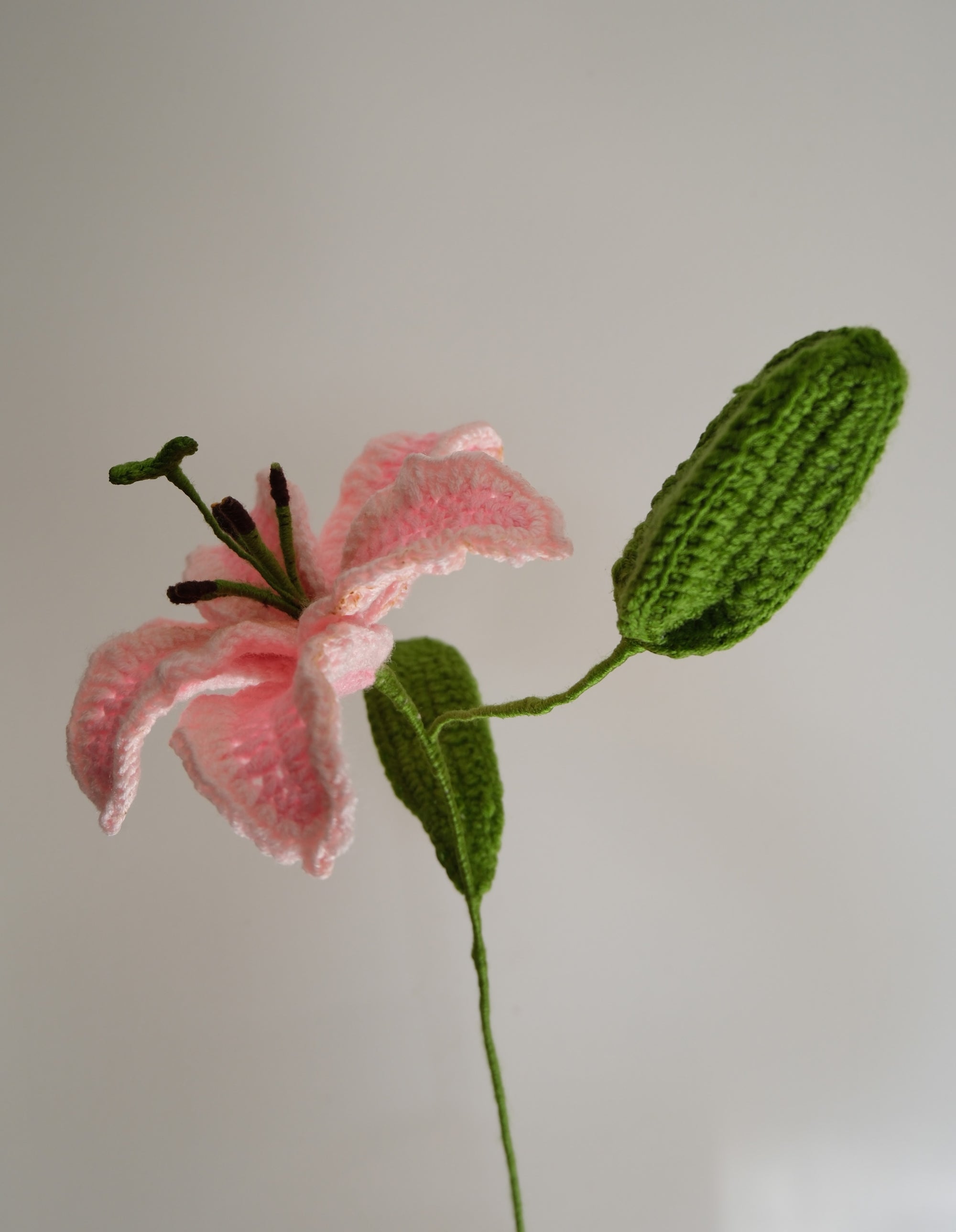 Handmade crochet stargazer lily in two-tone pink with one green leaf and an unopened flower bud, crafted with soft yarn on a green stem.