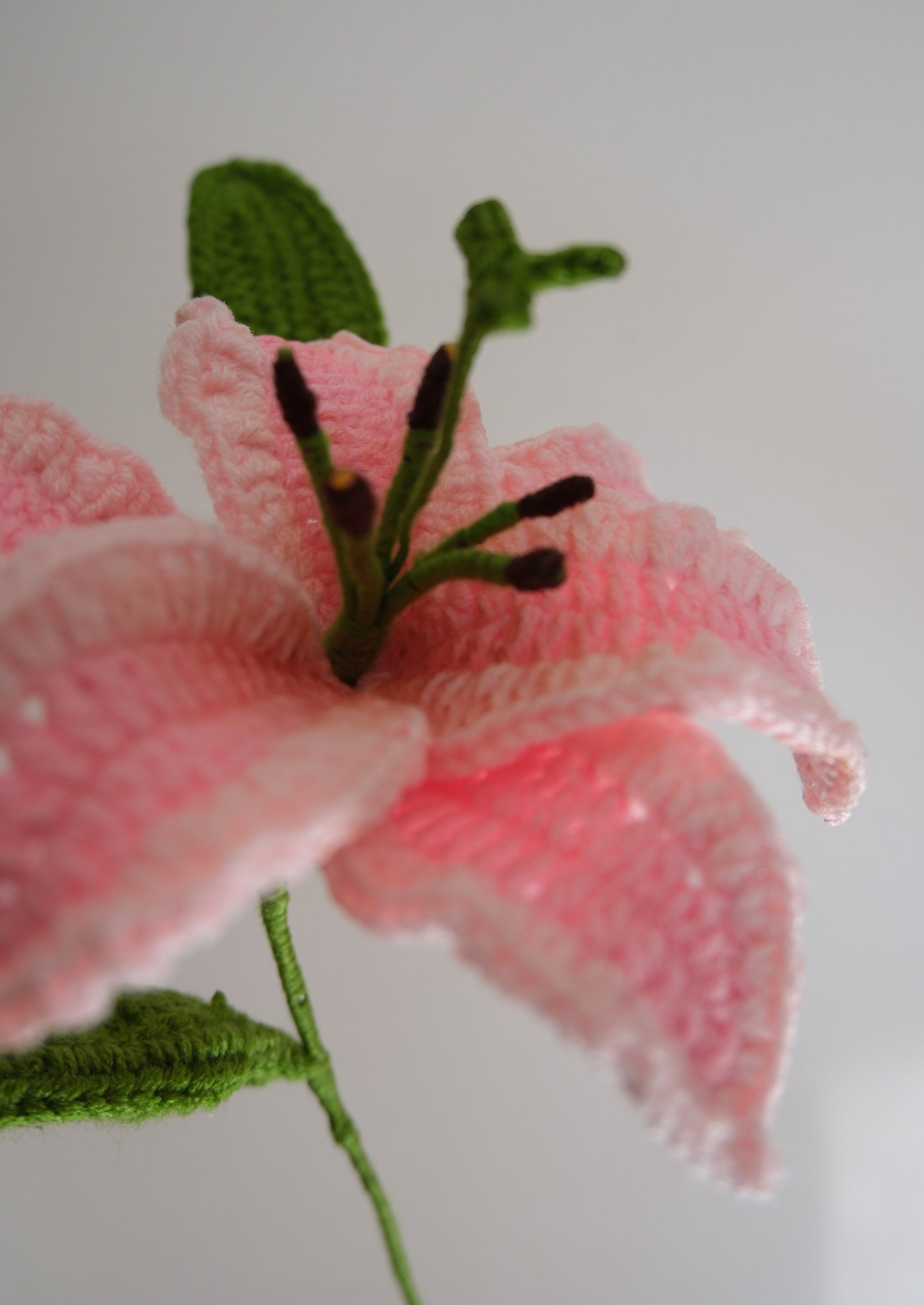 A close-up of a crocheted pink lily flower with green leaves and stem.