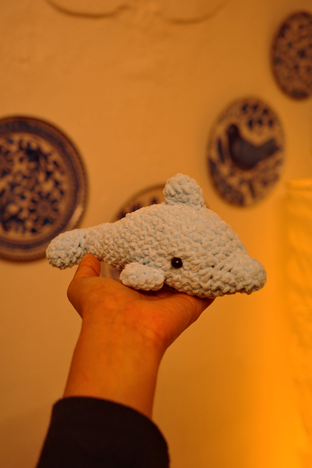 Hand holding a small plush dolphin amigurami toy against a yellow background with decorative patterns