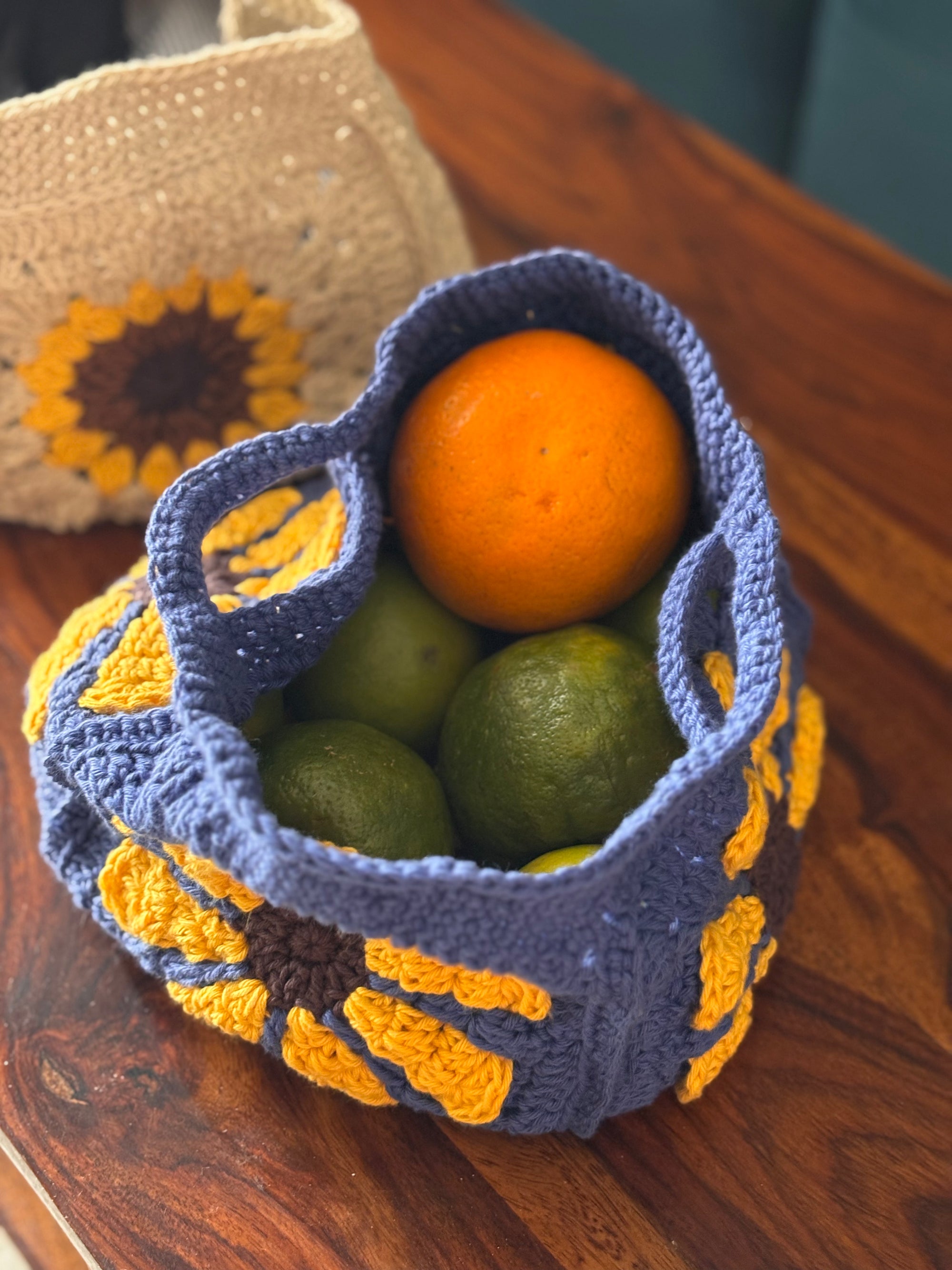 Blue crochet basket with sunflower patterns containing oranges and limes on a wooden surface.