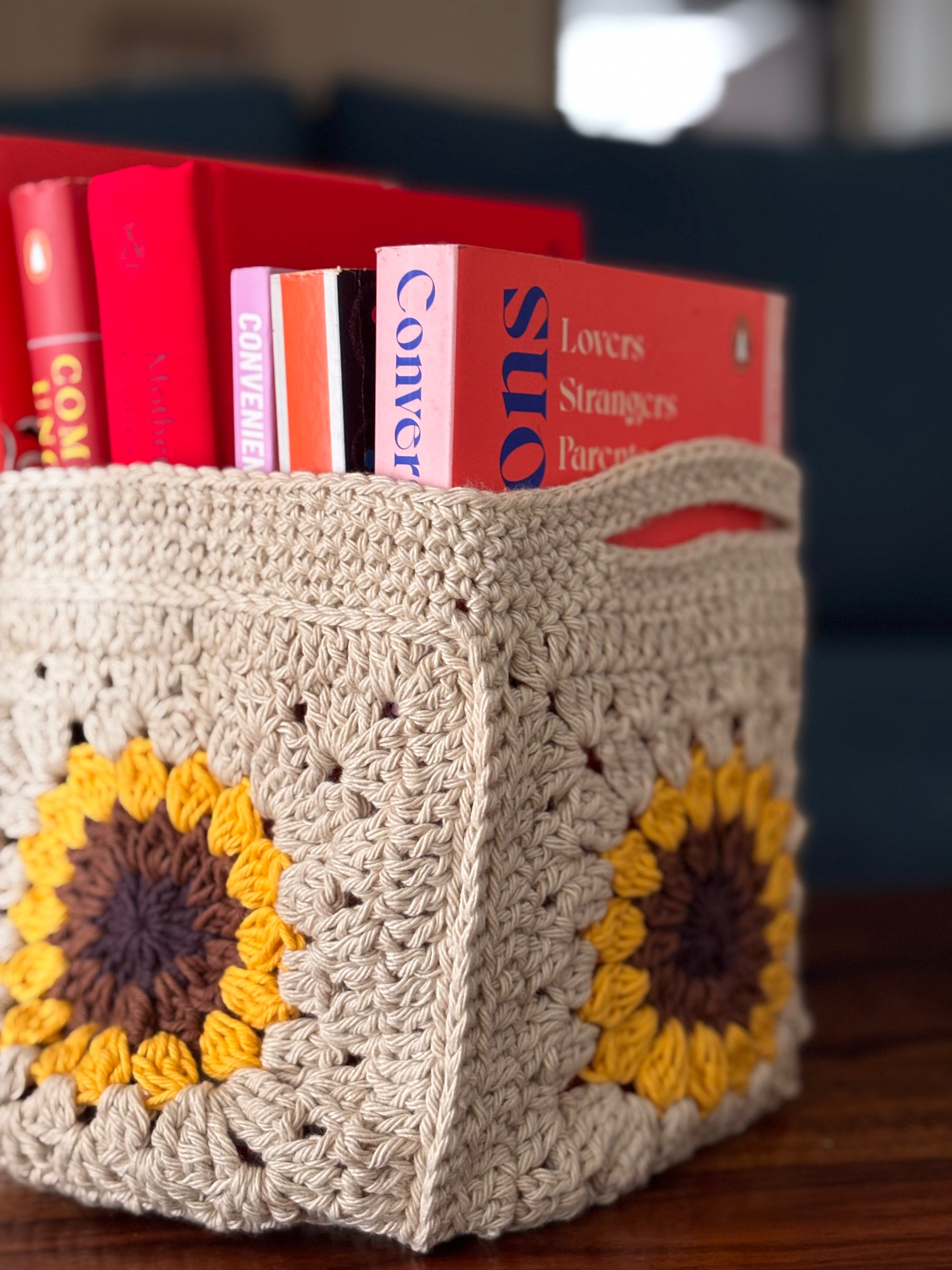 Crocheted basket with sunflower designs holding books on a wooden surface.