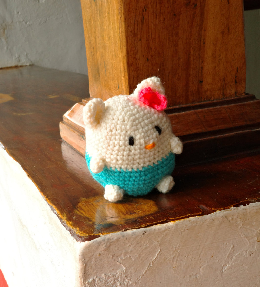 Small crocheted hello kitty toy with a teal sweater and red bow on a wooden surface