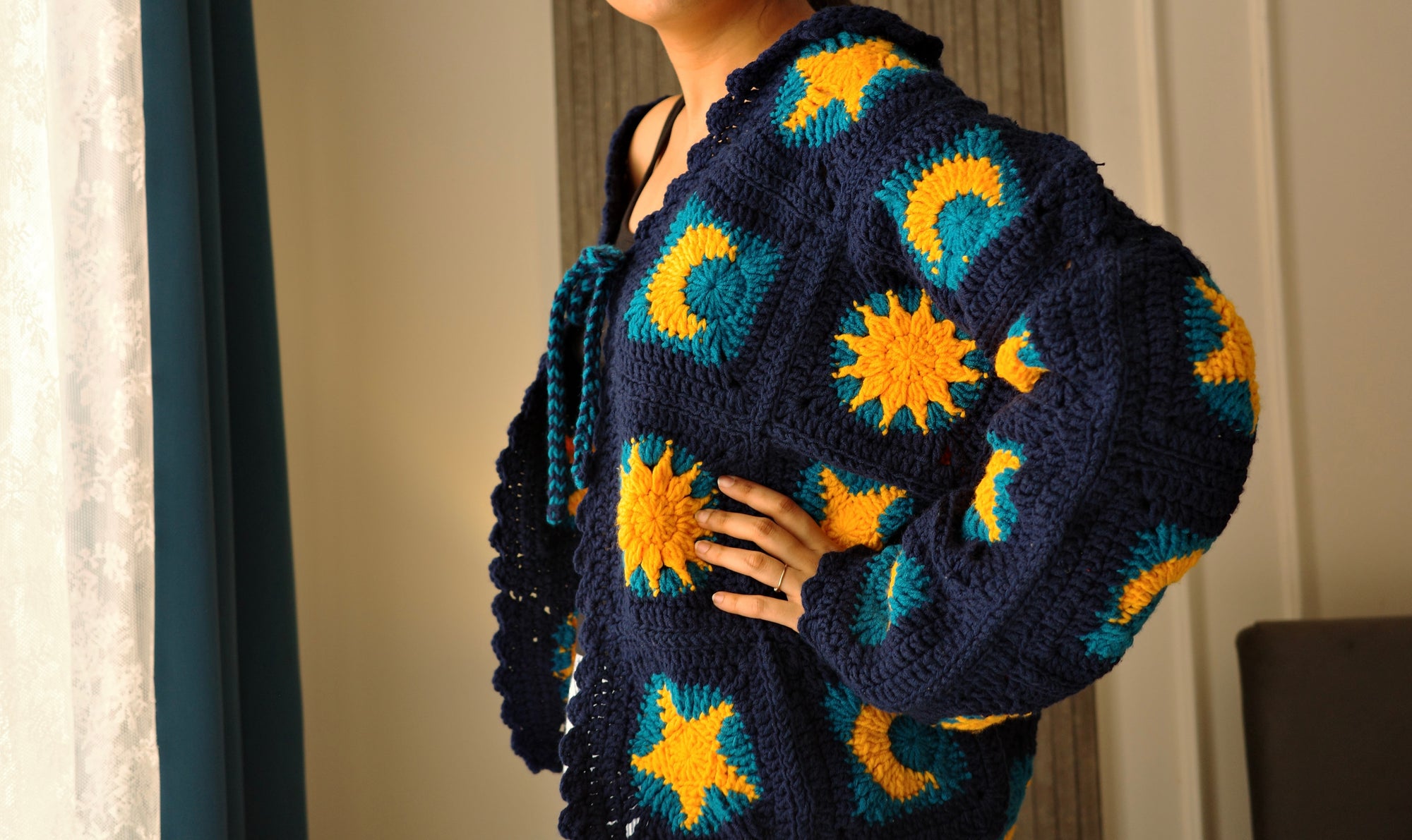 A person wearing a handmade crocheted cardigan with a navy blue base and yellow sunburst and teal crescent moon patterns, standing indoors near a window.