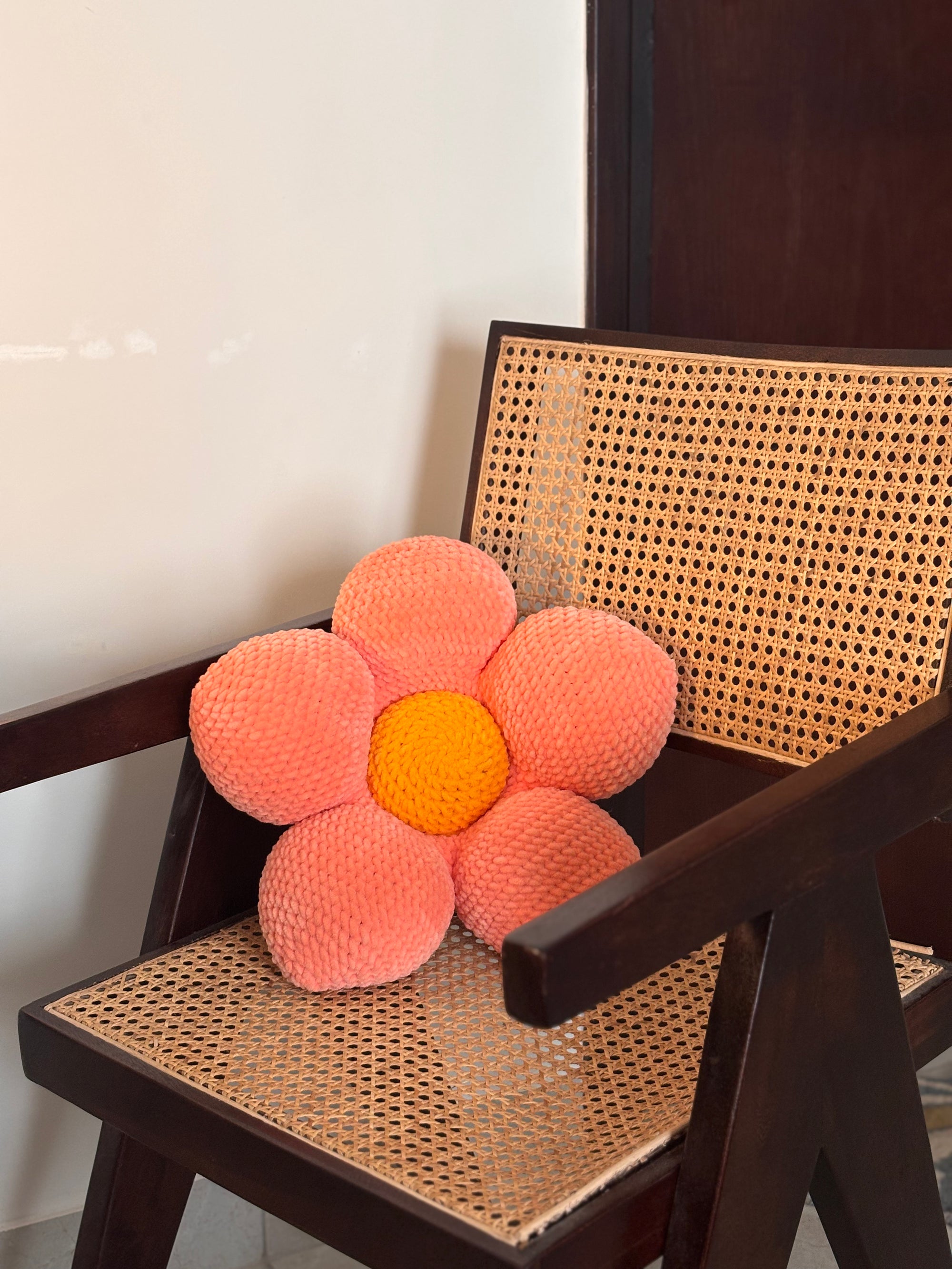 Flower-shaped crochet pink cushion with a yellow center on a wicker chair.
