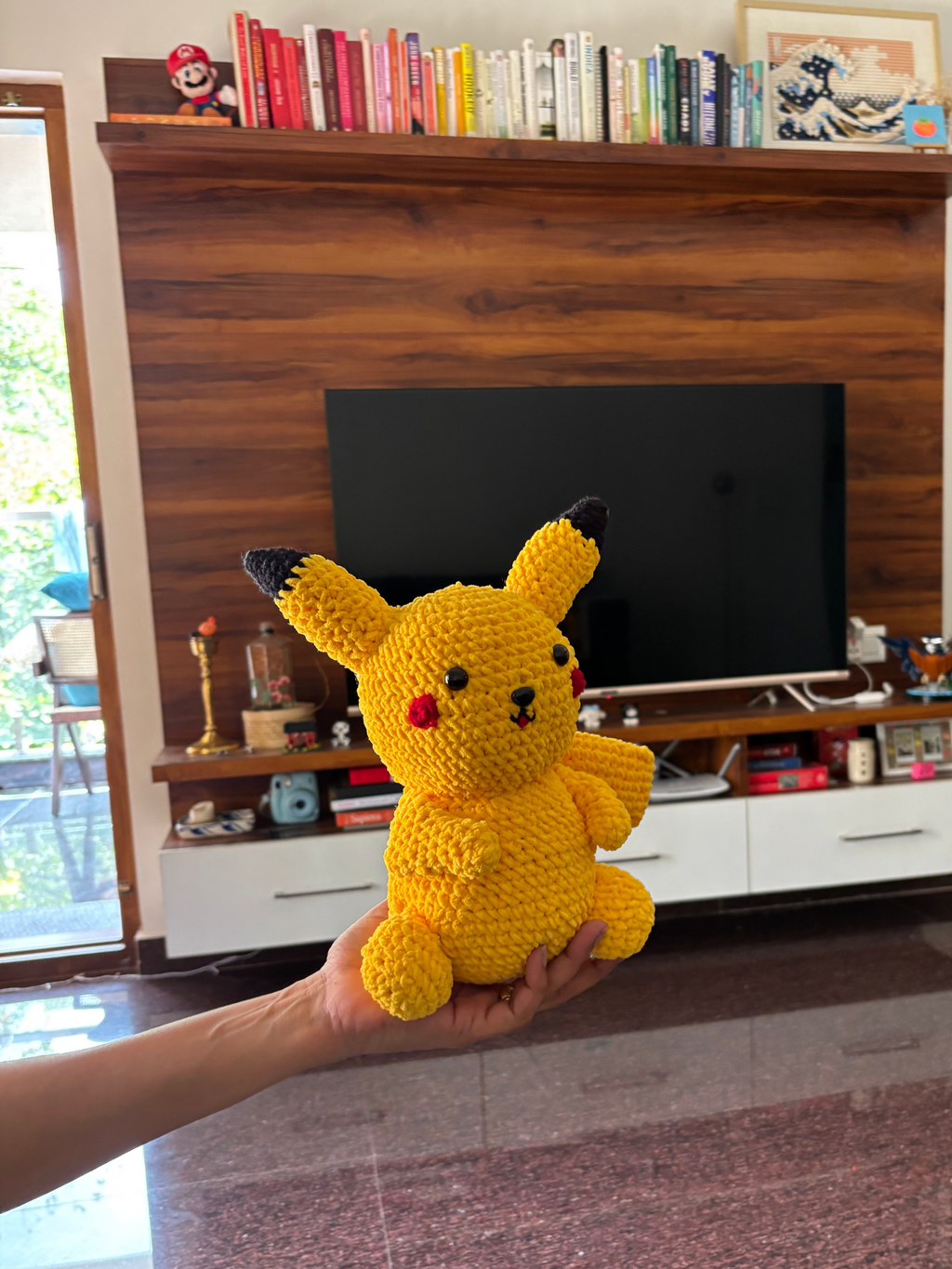Hand holding a yellow crochet Pikachu toy in a living room.