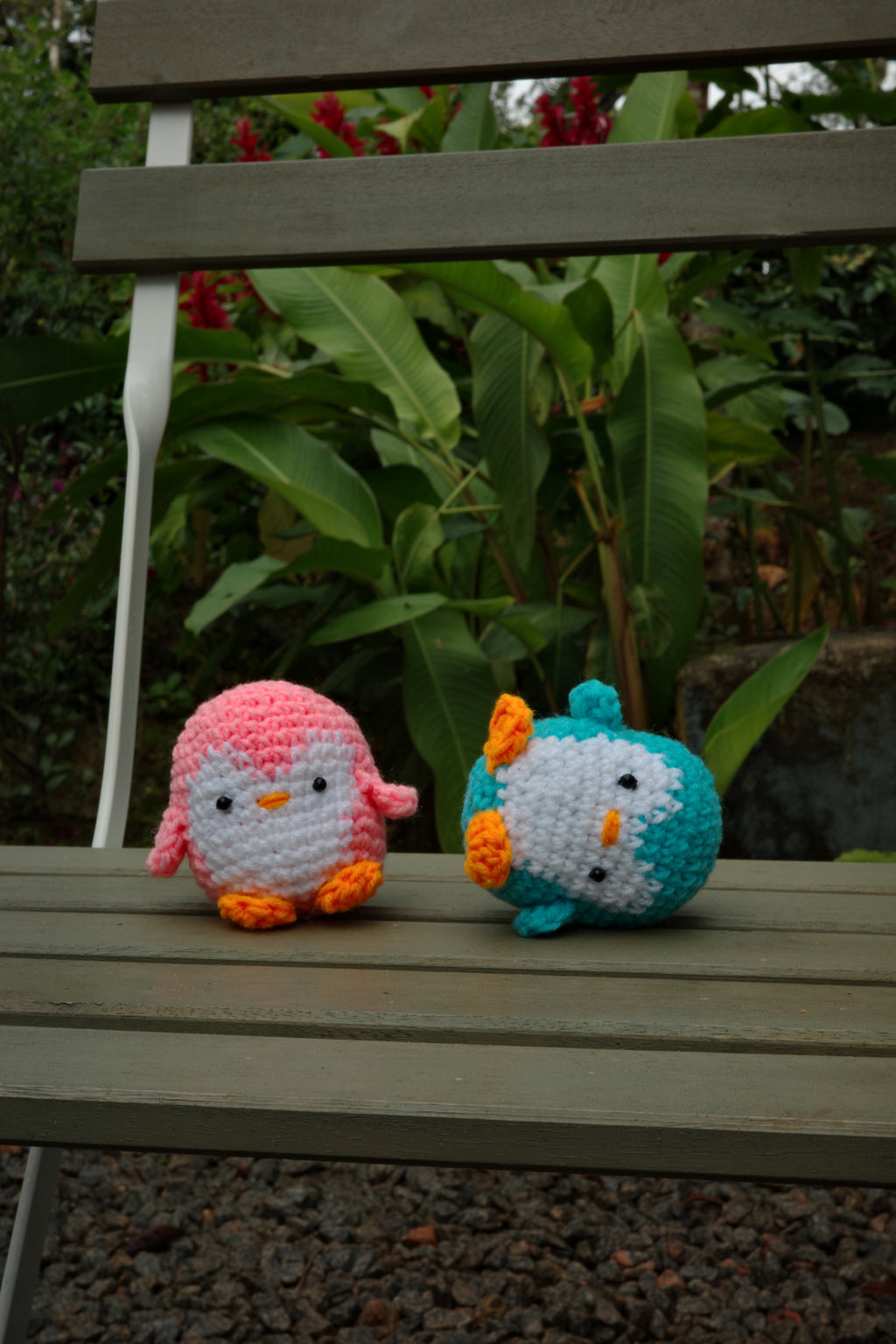 Two colorful crochet birds hanging on a wooden fence with greenery in the background.