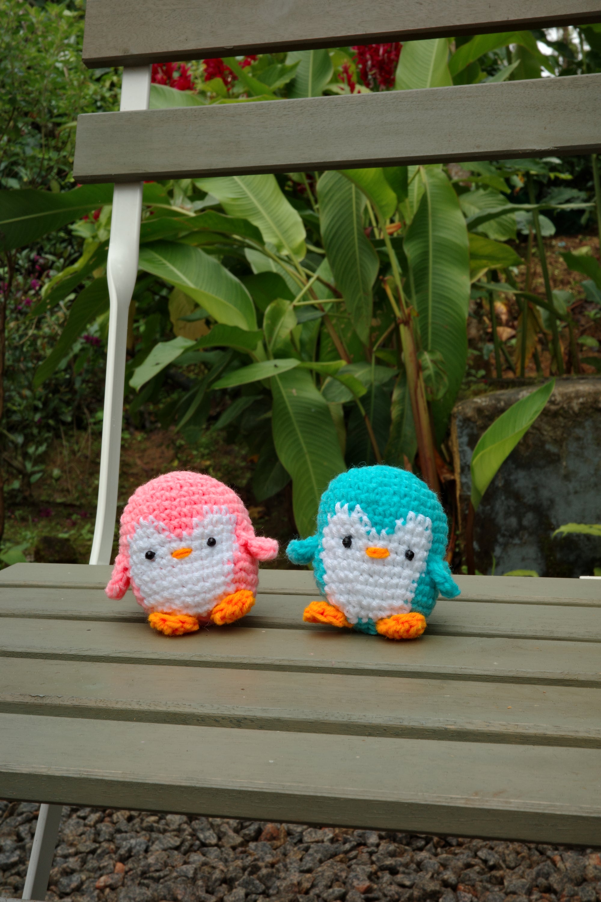Two knitted penguin toys hanging on a wooden fence with greenery in the background.