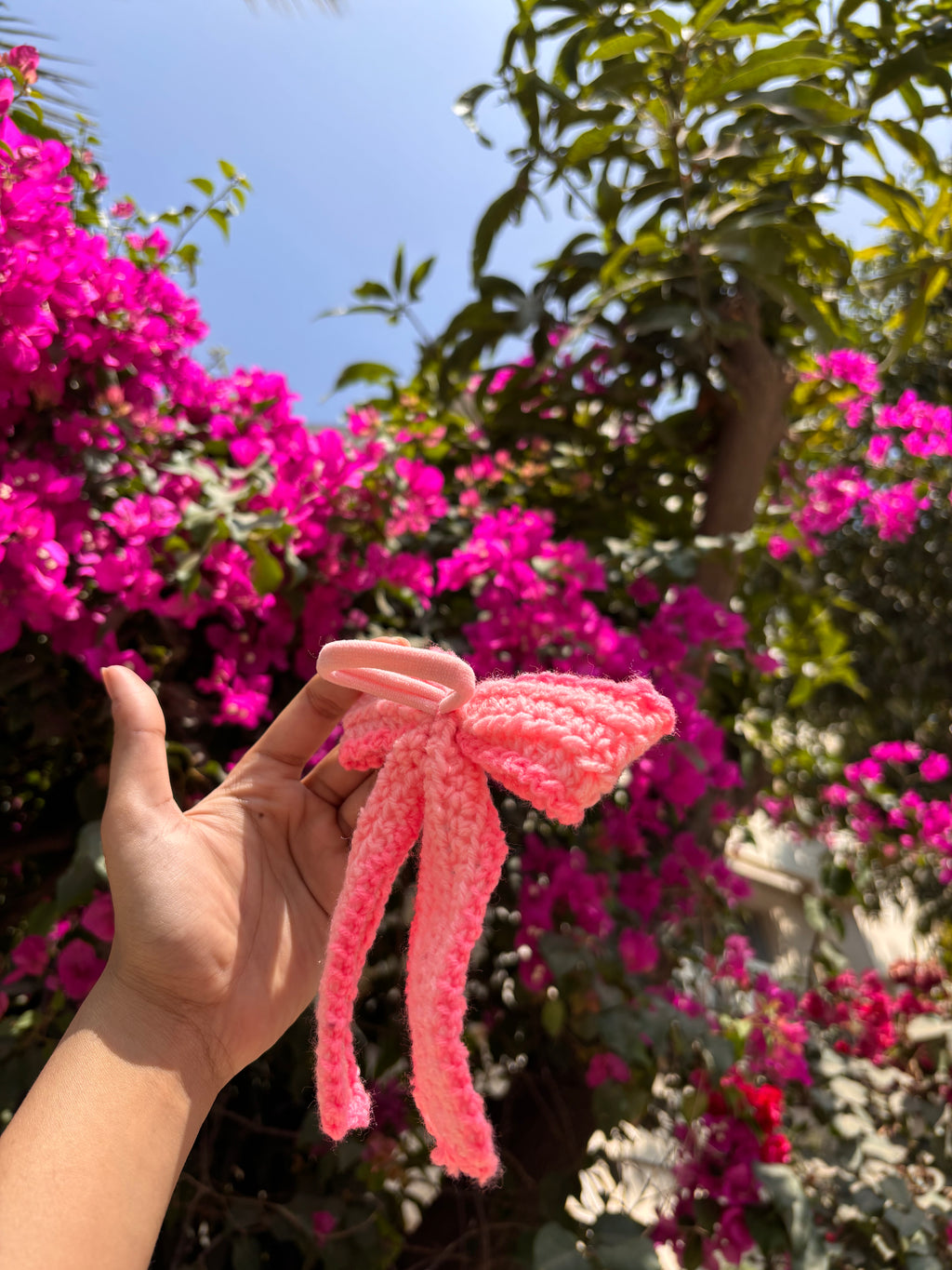 Hand holding a pink knitted bow with flowers in the background