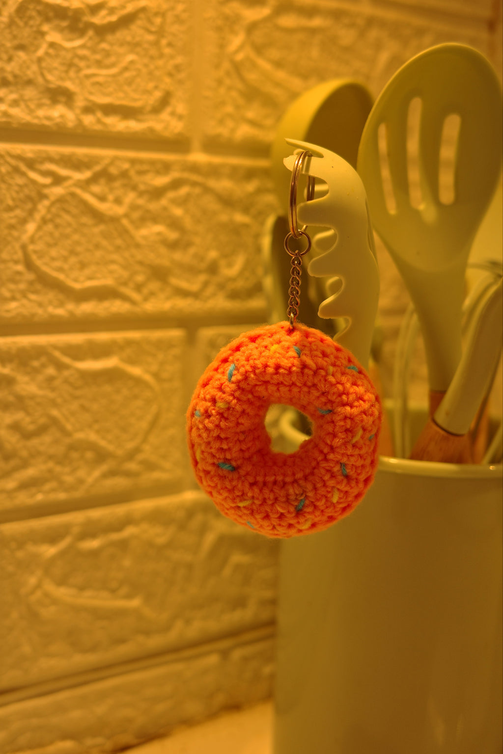 Crocheted donut with sprinkles on a yellow spoon against a textured wall.