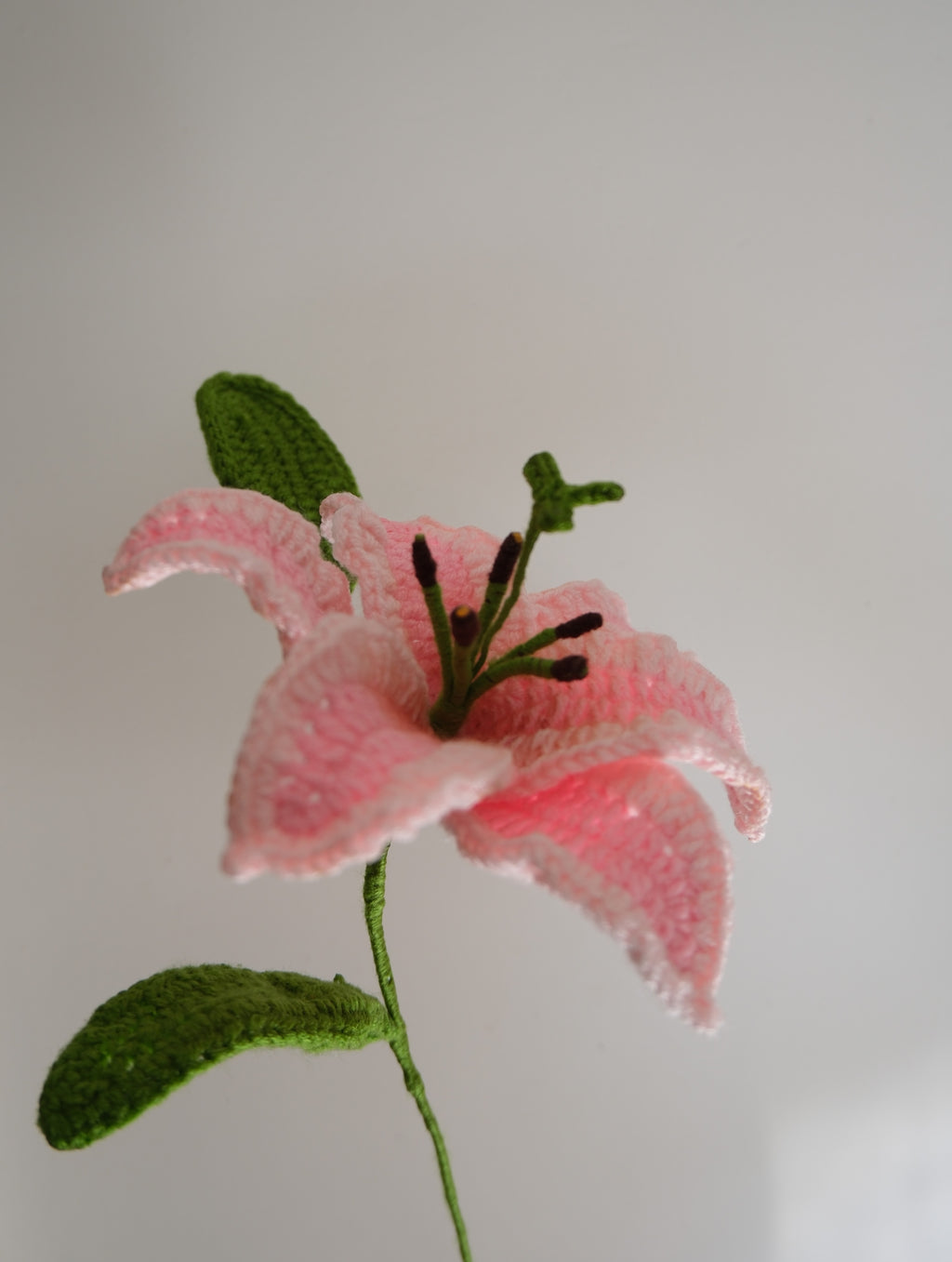 Handmade crochet stargazer lily in two-tone pink with one green leaf and an unopened flower bud, crafted with soft yarn on a green stem.