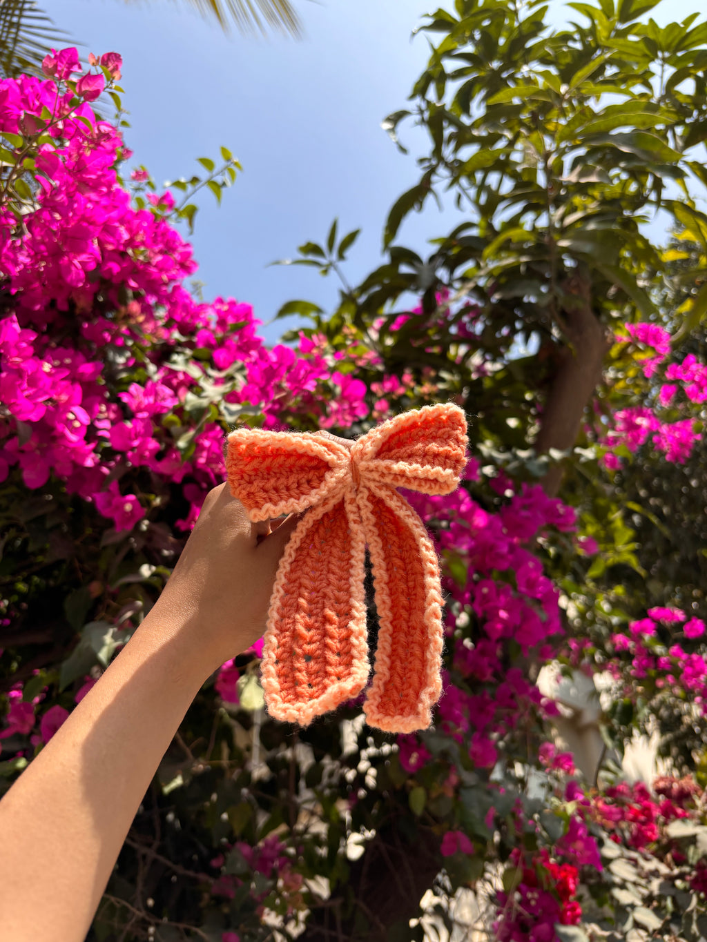 Hand holding a pink knitted bow with pink flowers and green leaves in the background