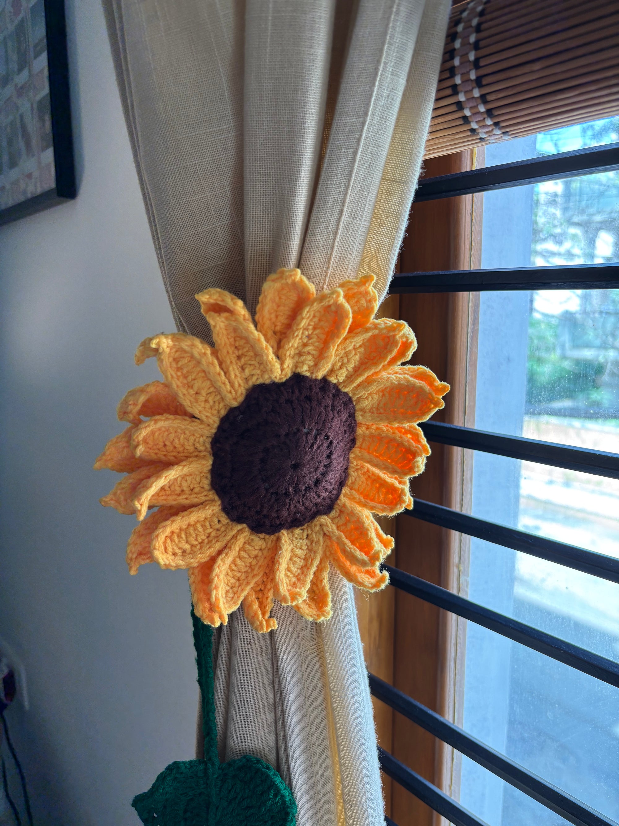 Crocheted sunflower curtain tieback attached to a beige curtain by a window.