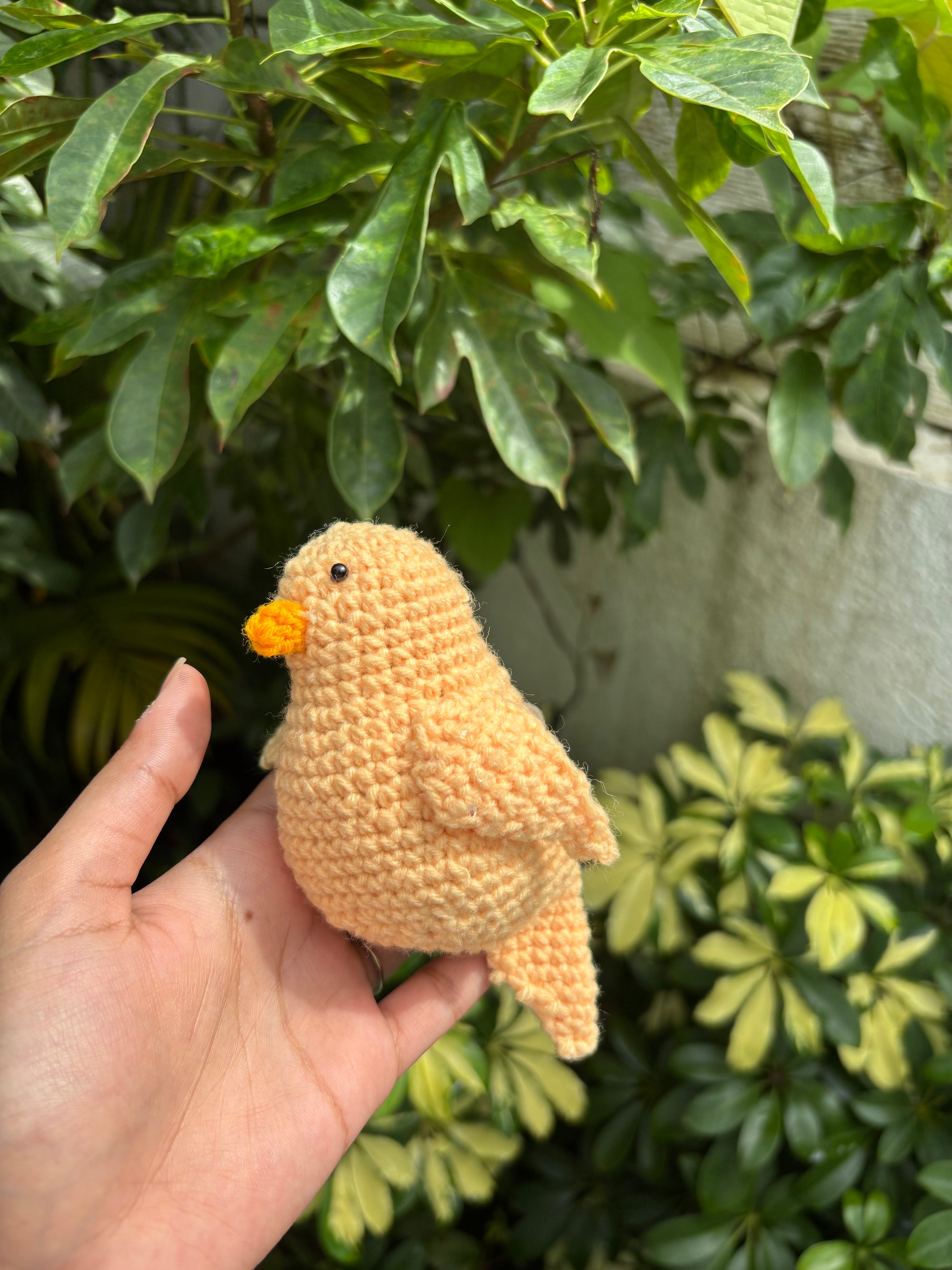 Hand holding a small crochet bird in front of green foliage