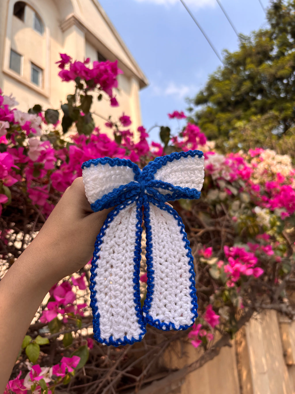 Hand holding a white and blue crocheted bow with flowers and a building in the background