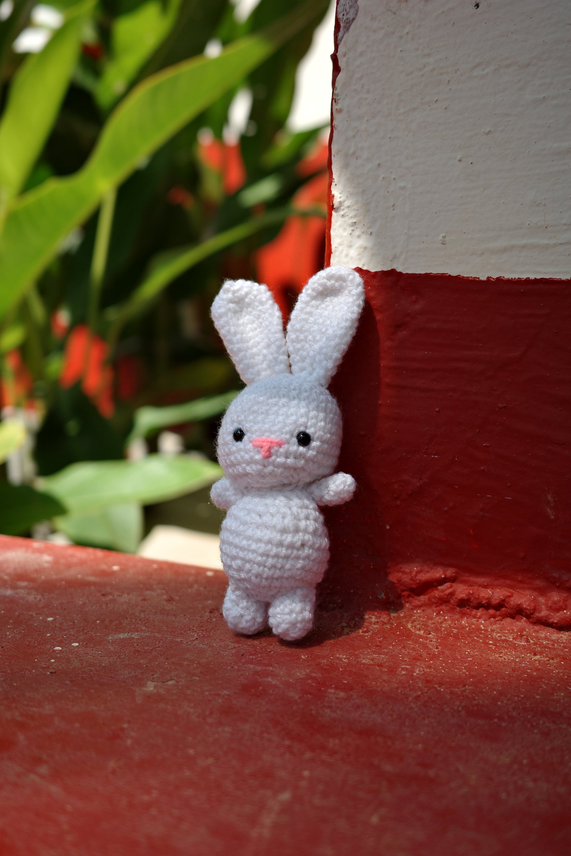 Small white crocheted bunny toy on a red surface with green foliage in the background