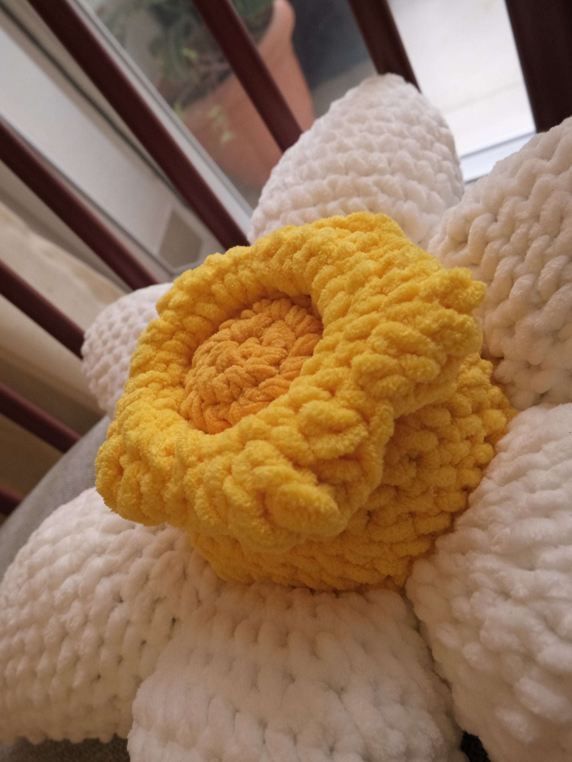 A close-up of a chunky crocheted flower pillow with a bright yellow textured center and large white petals.