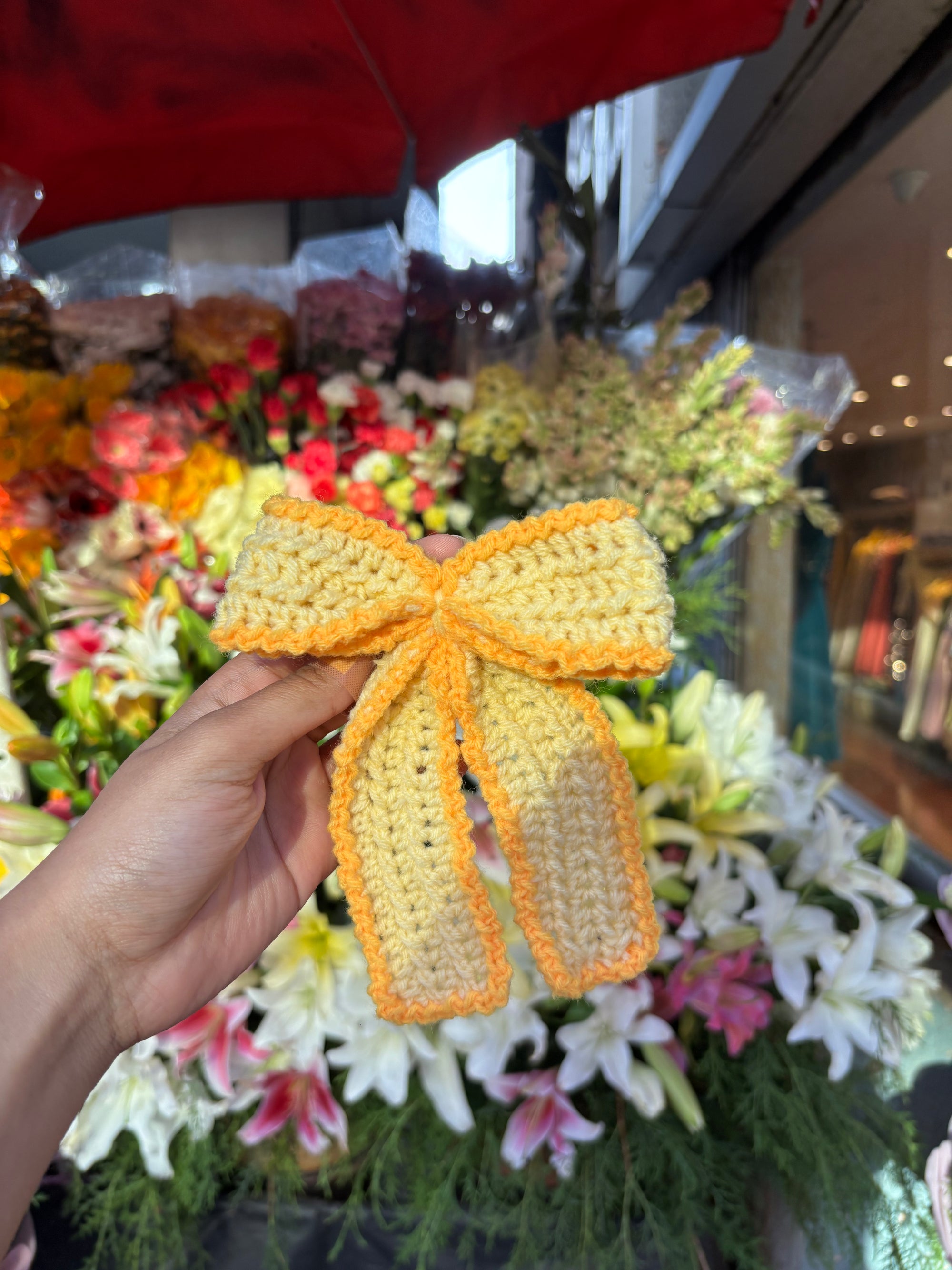 Hand holding a yellow and orange crocheted bow with floral arrangements in the background