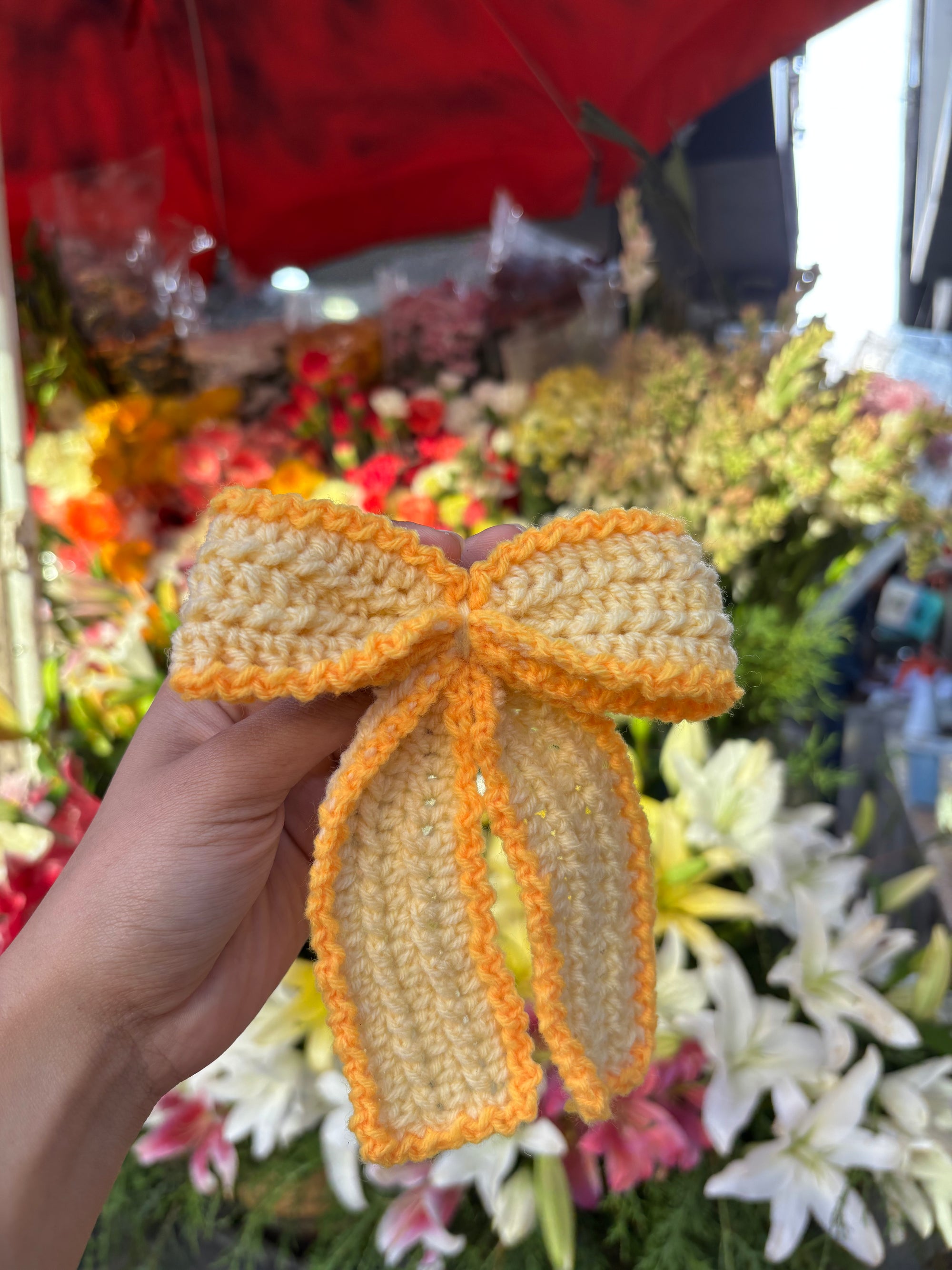 Hand holding a yellow and orange knitted bow with a floral background