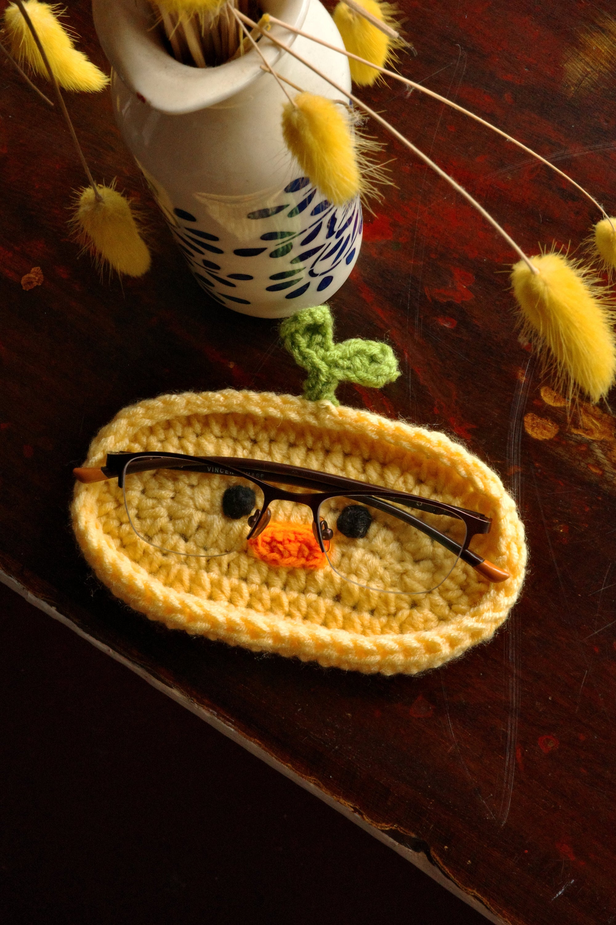 Crocheted eyeglass case with a face design on a wooden surface with dried plants.