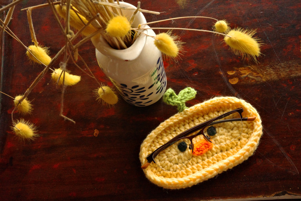Crocheted eye-shaped object with glasses on a wooden surface with dried plants.